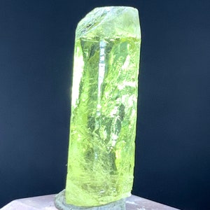 Puede incluir: Un cristal transl&uacute;cido amarillo brillante con una parte superior puntiaguda y una base plana. El cristal est&aacute; sentado sobre una superficie gris.