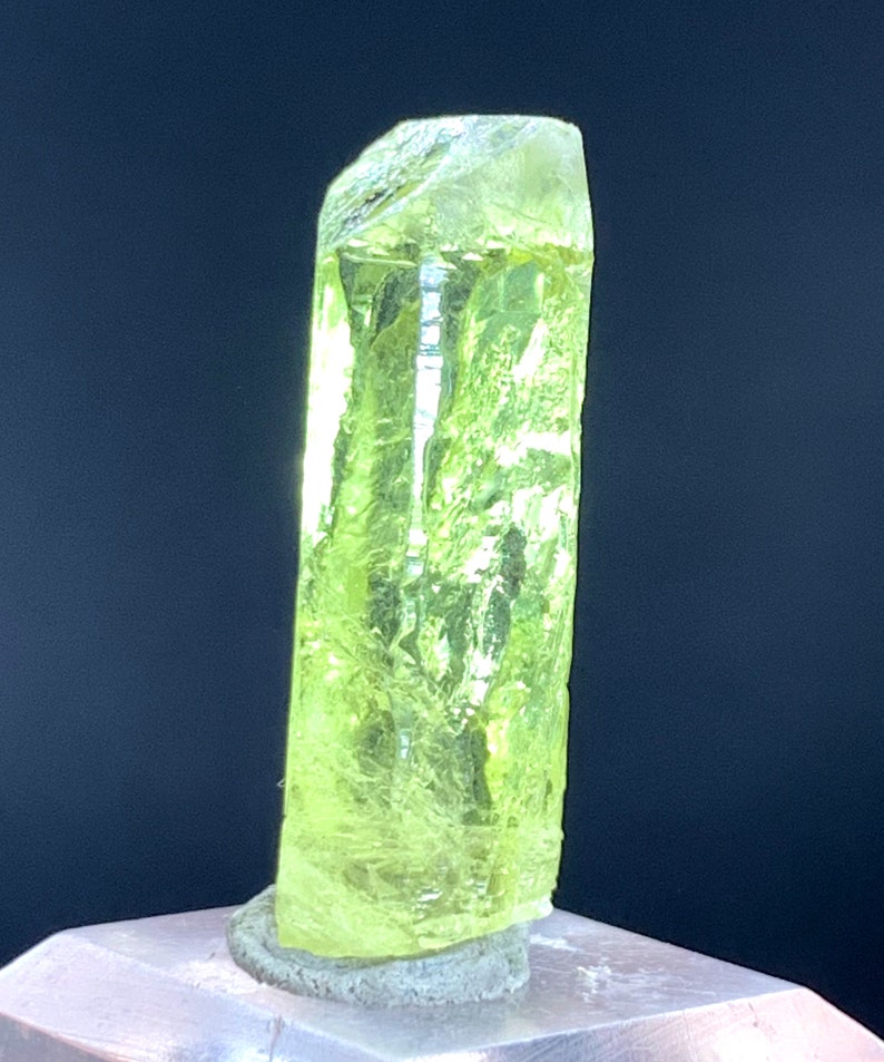 Puede incluir: Un cristal transl&uacute;cido amarillo brillante con una parte superior puntiaguda y una base plana. El cristal est&aacute; sentado sobre una superficie gris.