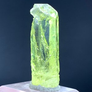 Puede incluir: Un cristal transl&uacute;cido amarillo brillante con una parte superior puntiaguda y una superficie rugosa y texturizada. El cristal est&aacute; montado sobre una base gris.