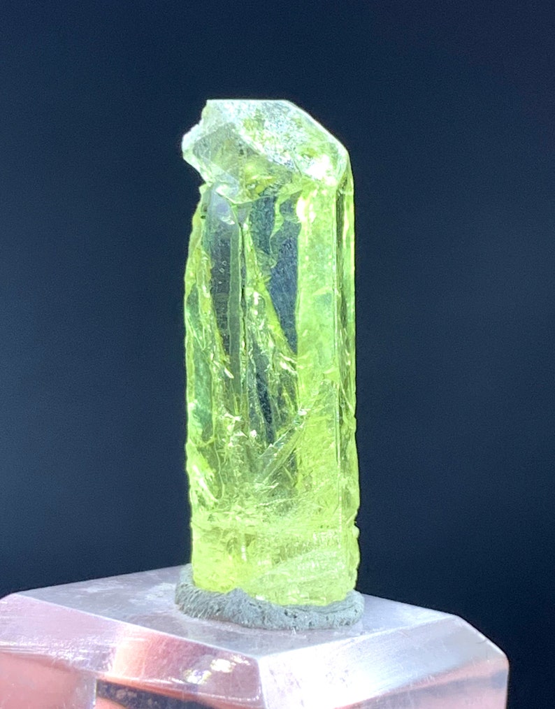 Puede incluir: Un cristal transl&uacute;cido amarillo brillante con una parte superior puntiaguda y una superficie rugosa y texturizada. El cristal est&aacute; montado sobre una base gris.