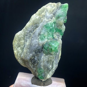 May include: A rough, gray rock with embedded green emeralds. The rock is on a white display stand.