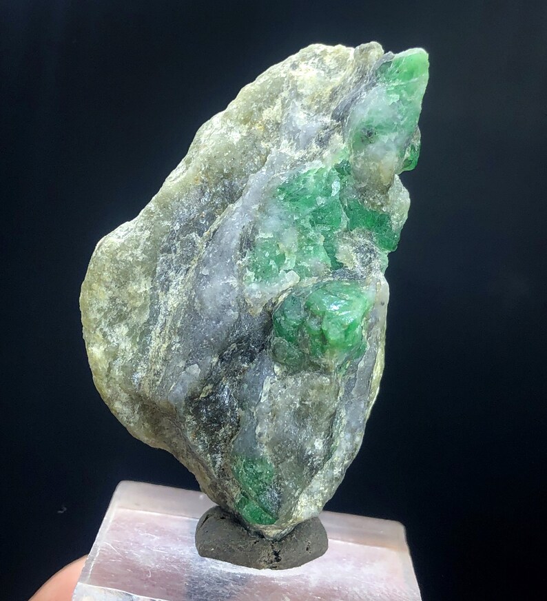 May include: A rough, gray rock with embedded green emeralds. The rock is on a white display stand.