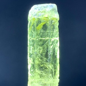 Puede incluir: Un cristal transl&uacute;cido verde brillante con una forma rectangular facetada. El cristal est&aacute; sobre una superficie blanca.