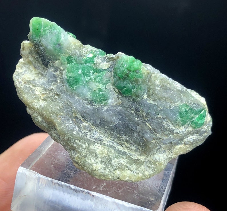 May include: A rough, gray rock with several bright green emerald crystals embedded in it. The rock is sitting on a clear display stand.