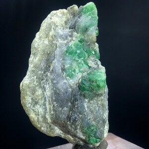 May include: A rough, gray rock with a cluster of bright green emeralds embedded in it. The rock is on a small, brown base.