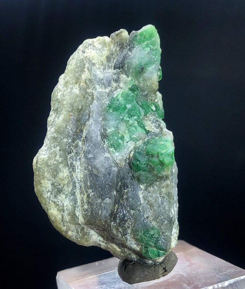 May include: A rough, gray rock with a cluster of bright green emeralds embedded in it. The rock is on a small, brown base.