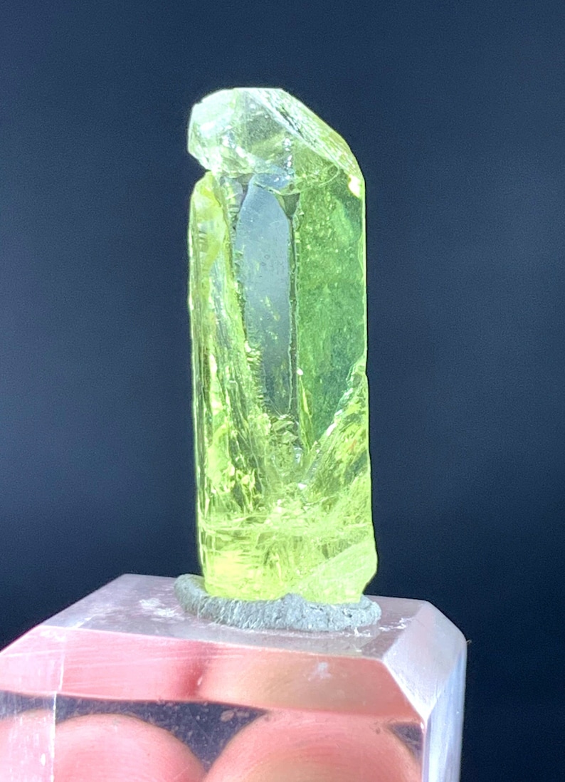 Puede incluir: Un cristal verde amarillo brillante con una textura clara y v&iacute;trea. El cristal est&aacute; sobre un soporte de exhibici&oacute;n de acr&iacute;lico transparente.