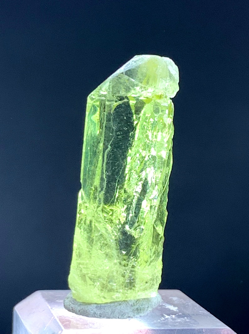 Puede incluir: Un gran cristal transl&uacute;cido verde brillante con una parte superior puntiaguda y una base plana. El cristal est&aacute; sentado sobre un pedestal blanco.