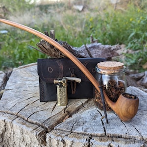 Puede incluir: Una pipa de fumar de madera con una boquilla larga y curva, una bolsa de cuero, un pequeño encendedor de latón y un frasco de vidrio lleno de tabaco.