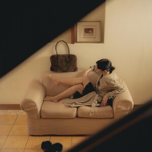 Peut inclure: Un canap&eacute; beige avec une personne portant un trench-coat beige et un short noir. Un sac &agrave; main en fourrure marron est pos&eacute; sur le canap&eacute;. Des pantoufles noires sont sur le sol carrel&eacute;. Une image encadr&eacute;e est accroch&eacute;e au mur.