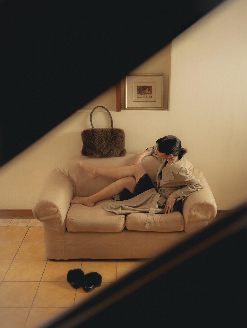 Peut inclure: Un canap&eacute; beige avec une personne portant un trench-coat beige et un short noir. Un sac &agrave; main en fourrure marron est pos&eacute; sur le canap&eacute;. Des pantoufles noires sont sur le sol carrel&eacute;. Une image encadr&eacute;e est accroch&eacute;e au mur.