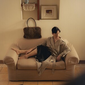 Peut inclure: Une personne portant un trench-coat beige et une jupe noire, assise sur un canap&eacute; beige. Un sac &agrave; main en fourrure marron repose sur le canap&eacute;. Des pantoufles en fourrure noire sont sur le sol. Un tableau encadr&eacute; et un vase avec des fleurs sont sur le mur.