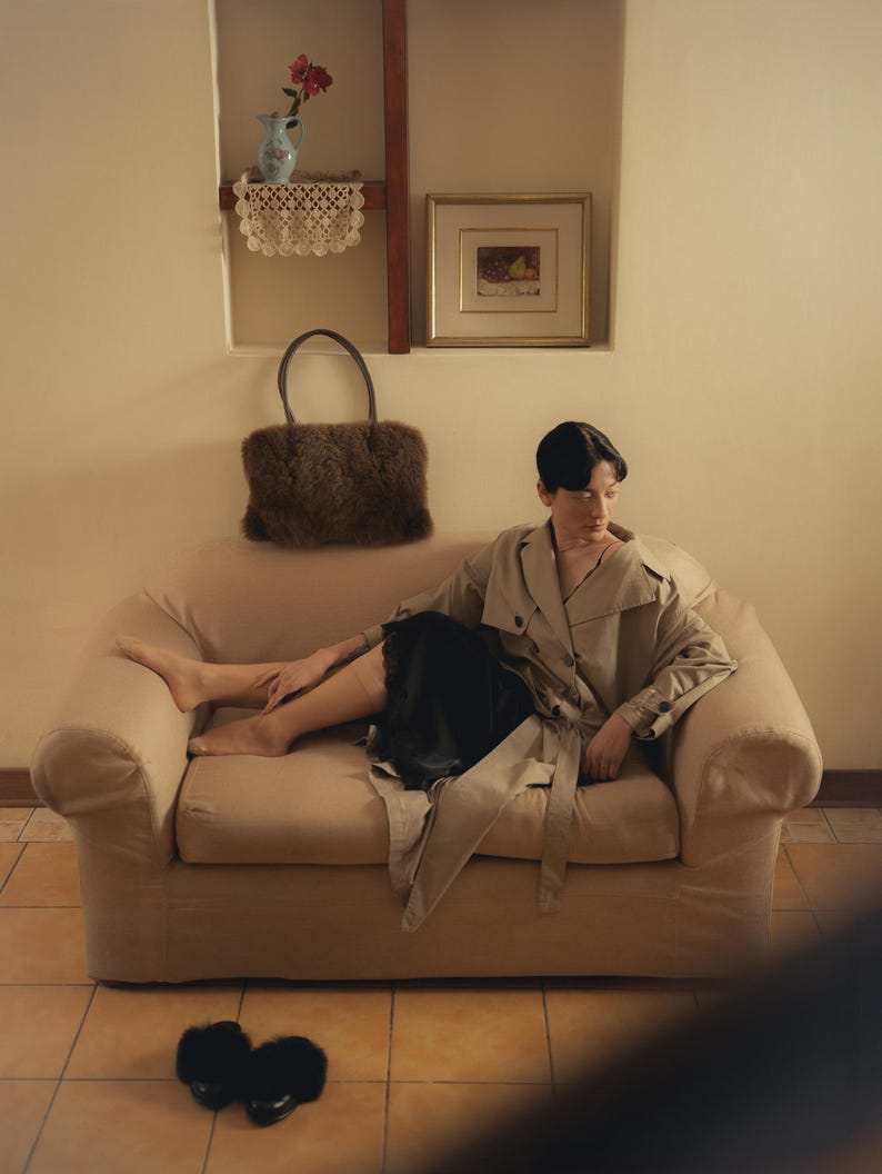 Peut inclure: Une personne portant un trench-coat beige et une jupe noire, assise sur un canap&eacute; beige. Un sac &agrave; main en fourrure marron repose sur le canap&eacute;. Des pantoufles en fourrure noire sont sur le sol. Un tableau encadr&eacute; et un vase avec des fleurs sont sur le mur.