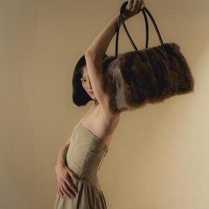 Peut inclure: Un sac &agrave; main en fausse fourrure marron avec des poign&eacute;es noires est lev&eacute;. Le sac a une forme rectangulaire et une fermeture &eacute;clair. Le mod&egrave;le porte une robe beige.