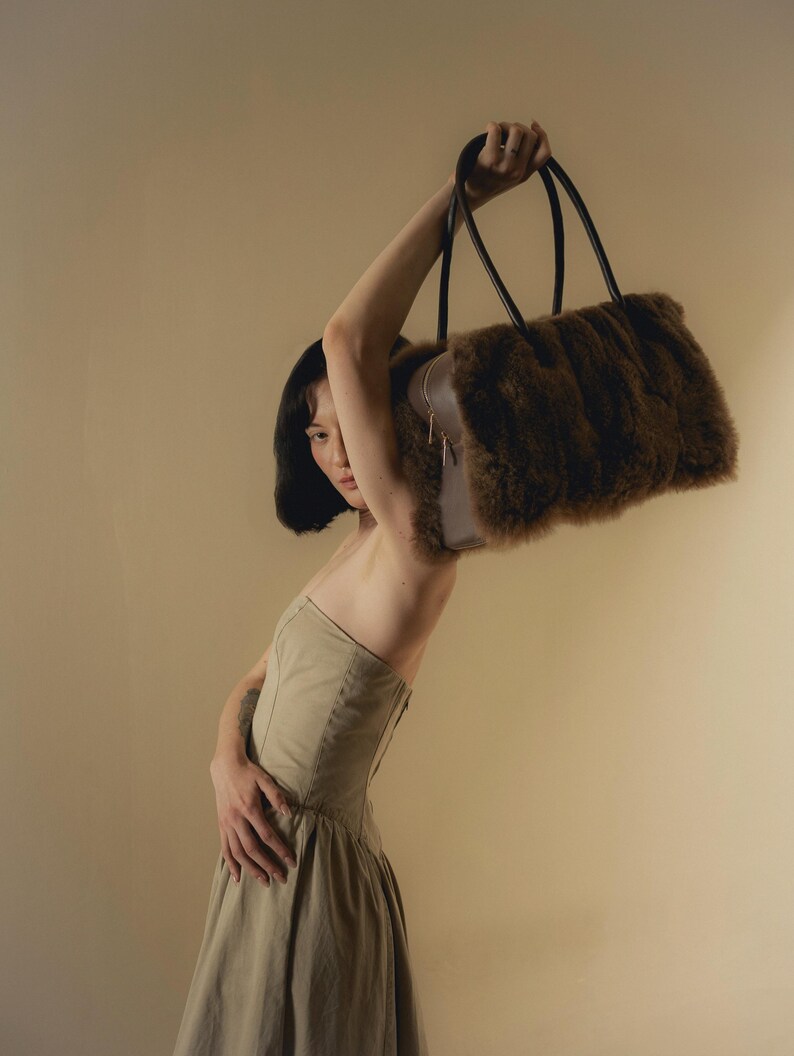 Peut inclure: Un sac &agrave; main en fausse fourrure marron avec des poign&eacute;es noires est lev&eacute;. Le sac a une forme rectangulaire et une fermeture &eacute;clair. Le mod&egrave;le porte une robe beige.