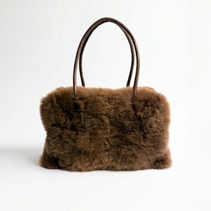 Peut inclure: Sac &agrave; main rectangulaire marron avec un ext&eacute;rieur duveteux et textur&eacute;. Le sac a deux poign&eacute;es arrondies marron fonc&eacute;. Le sac est pos&eacute; sur un fond blanc.