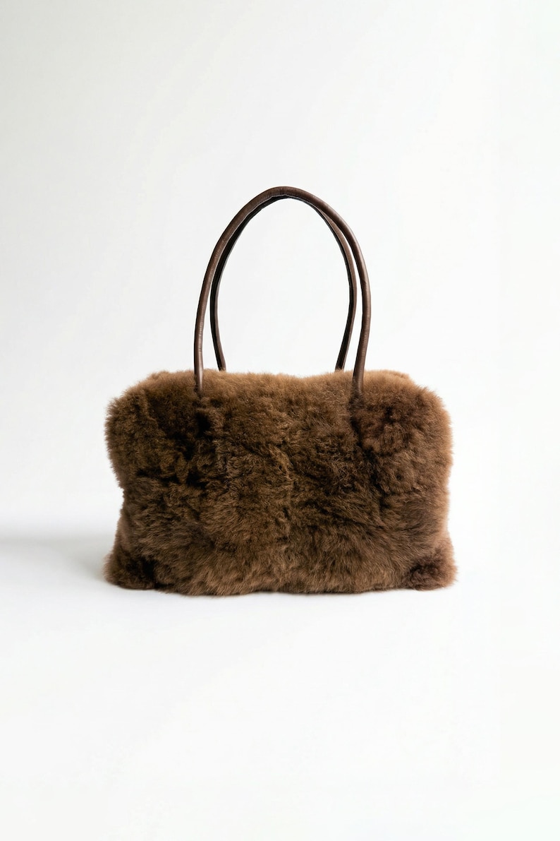Peut inclure: Sac &agrave; main rectangulaire marron avec un ext&eacute;rieur duveteux et textur&eacute;. Le sac a deux poign&eacute;es arrondies marron fonc&eacute;. Le sac est pos&eacute; sur un fond blanc.