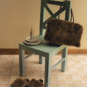 Peut inclure: Un sac fourre-tout en fausse fourrure marron avec des poign&eacute;es marron fonc&eacute; repose sur une chaise en bois bleu clair. Une paire de chaussons assortis en fausse fourrure marron sont au sol. Une figurine et des assiettes blanches sont sur la chaise.