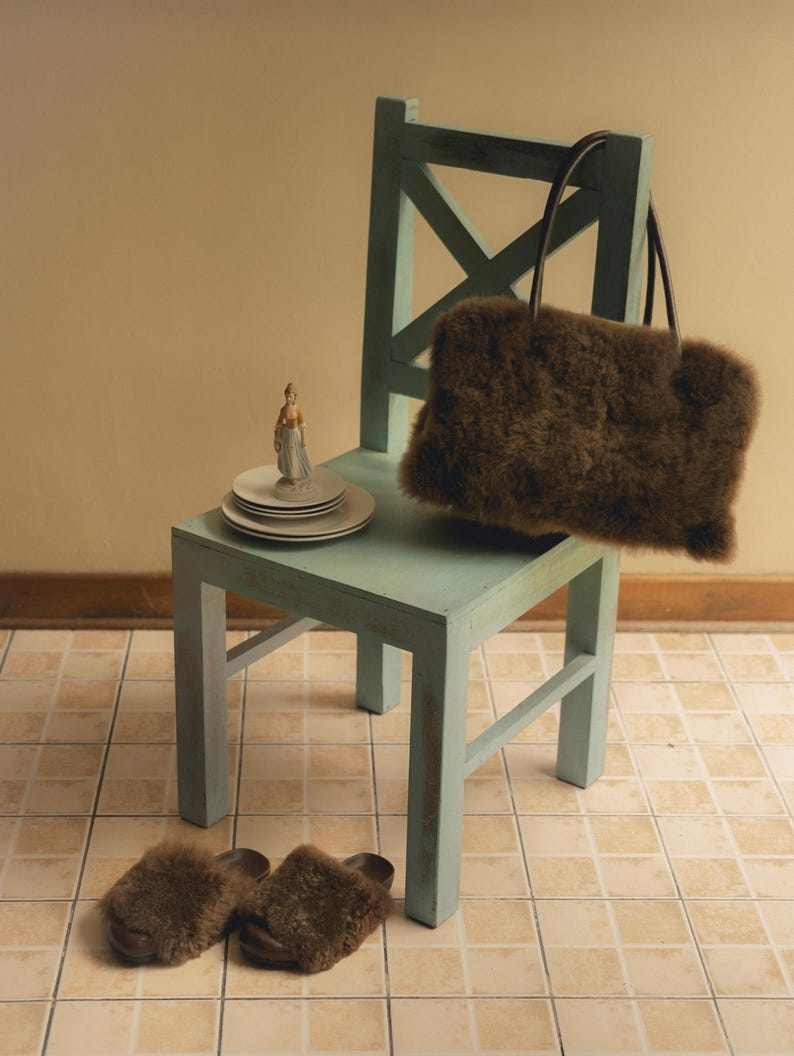 Peut inclure: Un sac fourre-tout en fausse fourrure marron avec des poign&eacute;es marron fonc&eacute; repose sur une chaise en bois bleu clair. Une paire de chaussons assortis en fausse fourrure marron sont au sol. Une figurine et des assiettes blanches sont sur la chaise.