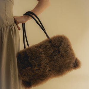 Peut inclure: Un sac &agrave; main rectangulaire marron avec un ext&eacute;rieur duveteux et textur&eacute;. Le sac a deux poign&eacute;es noires et arrondies. Le sac est tenu par une personne portant une robe de couleur claire.