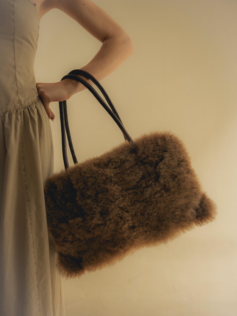 Peut inclure: Un sac &agrave; main rectangulaire marron avec un ext&eacute;rieur duveteux et textur&eacute;. Le sac a deux poign&eacute;es noires et arrondies. Le sac est tenu par une personne portant une robe de couleur claire.