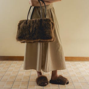 Peut inclure: Un sac fourre-tout marron et duveteux avec des poign&eacute;es noires est tenu devant une personne. Des pantoufles marron et duveteuses assorties sont port&eacute;es aux pieds. La personne porte une robe beige. Le sol est carrel&eacute;.
