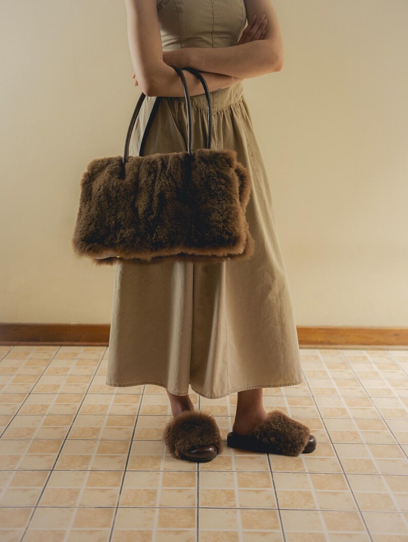 Peut inclure: Un sac fourre-tout marron et duveteux avec des poign&eacute;es noires est tenu devant une personne. Des pantoufles marron et duveteuses assorties sont port&eacute;es aux pieds. La personne porte une robe beige. Le sol est carrel&eacute;.