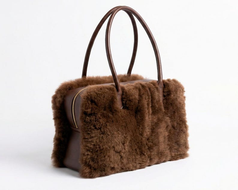 Peut inclure: Un sac &agrave; main marron de forme rectangulaire avec deux anses arqu&eacute;es. Le sac pr&eacute;sente un ext&eacute;rieur duveteux et textur&eacute; et une base en cuir marron. Une fermeture &eacute;clair dor&eacute;e traverse le dessus.