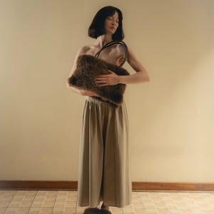 Peut inclure: Un sac &agrave; main marron et duveteux avec une poign&eacute;e fonc&eacute;e est tenu devant une personne. La personne porte un pantalon marron &agrave; jambes larges et des pantoufles moelleuses assorties. L'arri&egrave;re-plan est un mur beige clair et un sol carrel&eacute;.
