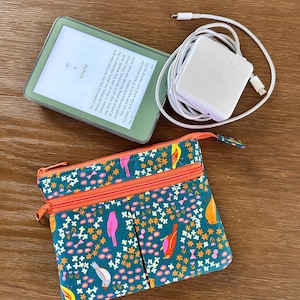 May include: A light green e-reader displaying text, a white power adapter with a coiled charging cable, and a teal fabric pouch featuring a colorful pattern of small birds and flowers with two orange zippered compartments.