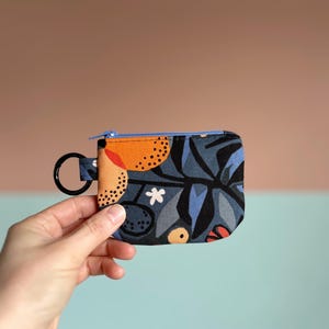 May include: A small, patterned coin purse with a blue zipper and a black key ring. The fabric features a dark blue background with orange, white, and blue floral and abstract shapes. The purse is held against a pink and blue background.