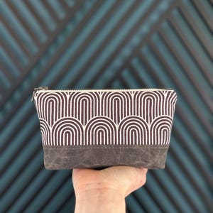 May include: A rectangular zippered pouch featuring a dark brown and white geometric arch pattern on the upper portion and a solid dark gray base. The pouch has a silver-toned metal zipper closure along the top edge and a structured fabric construction.