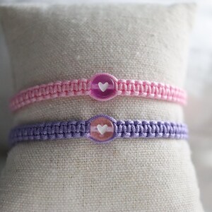 May include: Two macrame bracelets, one pink and one purple, each with a heart-shaped charm. The bracelets are displayed on a beige fabric surface. The charms are clear with a white heart.