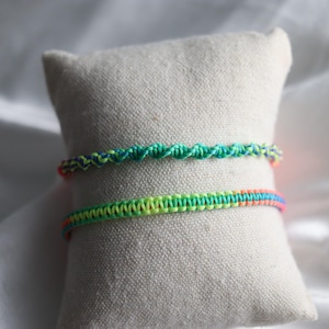 May include: Two handmade macrame bracelets in vibrant colors. The top bracelet features a twisted design with green and blue threads. The bottom bracelet is a straight design with a gradient of yellow, green, orange, and pink. Both are displayed on a beige pillow.