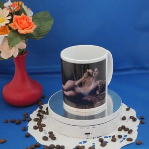 Könnte beinhalten: Weißer Keramikbecher mit einem Schwarz-Weiß-Bild einer nackten Person, die Blumen hält. Der Becher steht auf einem weißen Drehständer, umgeben von Kaffeebohnen. Eine rote Vase mit Blumen befindet sich im Hintergrund.