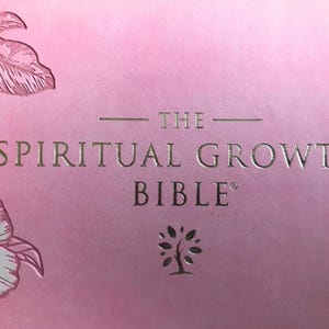 A pink book cover with the words "THE SPIRITUAL GROWTH BIBLE" in gold lettering. Floral illustrations in white and pink are in the upper left corner. A tree icon is below the word "BIBLE".