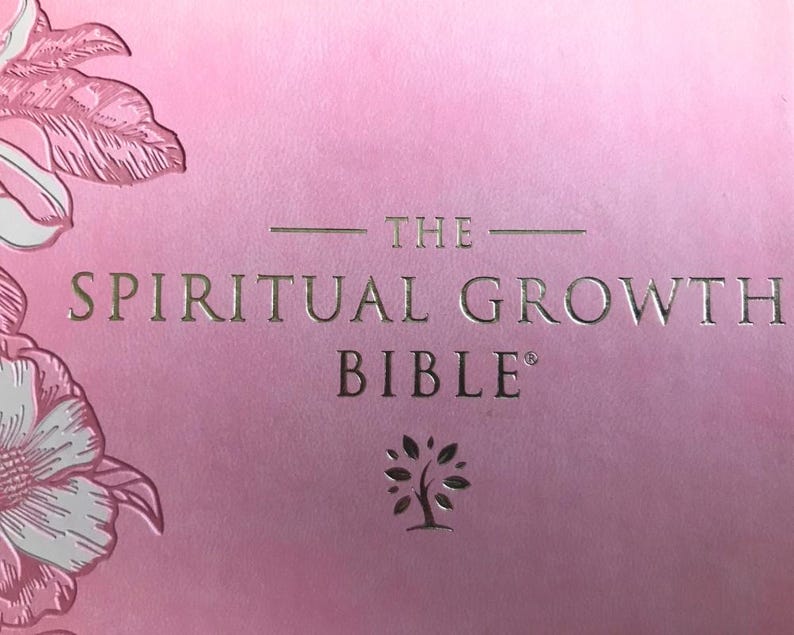 A pink book cover with the words "THE SPIRITUAL GROWTH BIBLE" in gold lettering. Floral illustrations in white and pink are in the upper left corner. A tree icon is below the word "BIBLE".