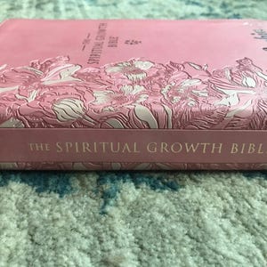 A pink Bible with embossed floral designs and the title "The Spiritual Growth Bible." The book's cover features a textured pattern and the word "Scripture" in a cursive font.