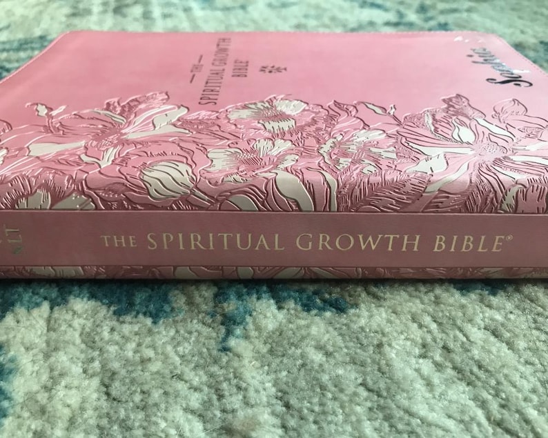 A pink Bible with embossed floral designs and the title "The Spiritual Growth Bible." The book's cover features a textured pattern and the word "Scripture" in a cursive font.