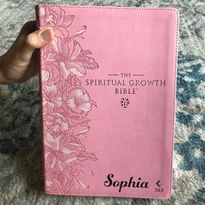 A pink Bible with the text "The Spiritual Growth Bible" and the name "Sophia" printed on the cover. Floral designs in a lighter pink adorn the upper left corner. The book is held by a hand.