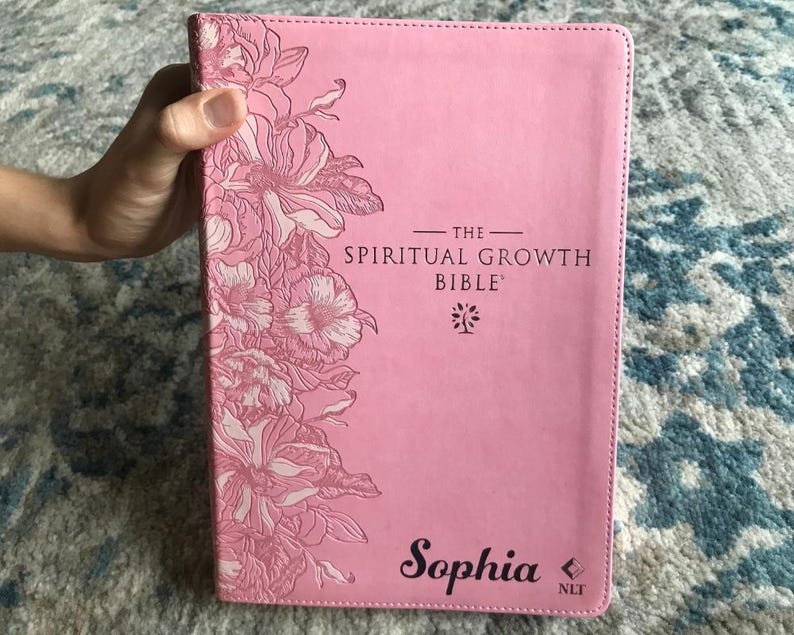 A pink Bible with the text "The Spiritual Growth Bible" and the name "Sophia" printed on the cover. Floral designs in a lighter pink adorn the upper left corner. The book is held by a hand.