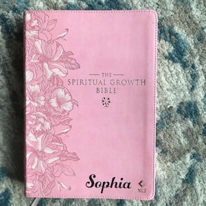 Pink Bible titled "The Spiritual Growth Bible" with floral design and the name "Sophia". The cover features a stitched border and the letters "NLT".
