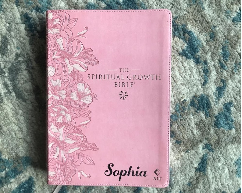Pink Bible titled "The Spiritual Growth Bible" with floral design and the name "Sophia". The cover features a stitched border and the letters "NLT".