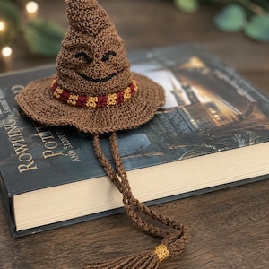 May include: A brown crocheted hat with a pointed top and a wide brim, featuring a stitched black smiling face. The hat has a maroon and gold patterned band around the base and two long brown crocheted cords ending in a brown tassel with a gold ring.