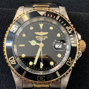 May include: A stainless steel Invicta watch with a black dial, gold accents, and a date display at the 3 o'clock position. The watch features a black rotating bezel with gold markings and a gold crown. The watch band is stainless steel.