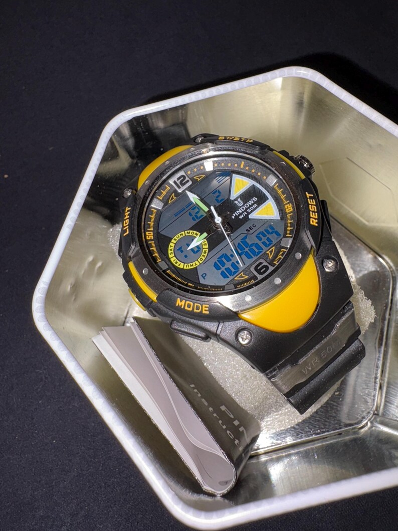Op de afbeelding: Een zwart en geel digitaal polshorloge met een zwarte band, in een zilveren metalen container. De wijzerplaat geeft de tijd en dag weer, met het woord "PINDOWS" zichtbaar. Het horloge heeft ook de woorden "LIGHT", "MODE" en "RESET".