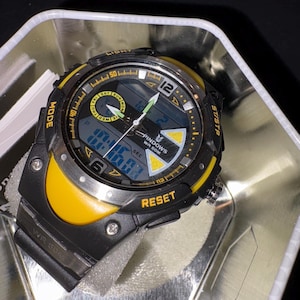 Op de afbeelding: Een zwart en geel digitaal polshorloge met een zwarte band. De wijzerplaat toont de tijd en dag, met de merknaam "PINDOWS" zichtbaar. Het horloge zit in een zilveren metalen container.