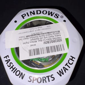 Op de afbeelding: Een witte, zeshoekige horlogedoos met een groene en zwarte rand. Op de doos staat "FASHION SPORTS WATCH" in zwarte tekst. Een label op de doos luidt "PINDOWS Watch for Men, Military Digital" en bevat een barcode.