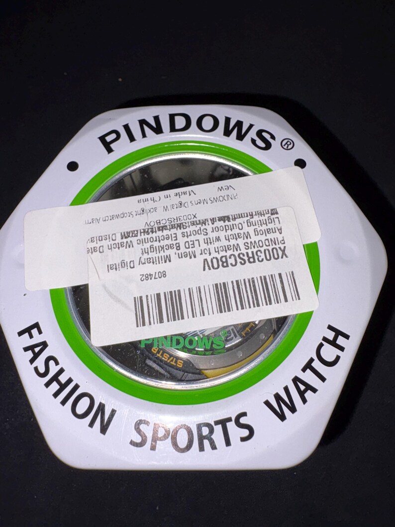 Op de afbeelding: Een witte, zeshoekige horlogedoos met een groene en zwarte rand. Op de doos staat "FASHION SPORTS WATCH" in zwarte tekst. Een label op de doos luidt "PINDOWS Watch for Men, Military Digital" en bevat een barcode.