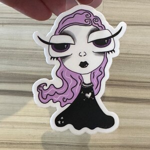 Peut inclure: Un autocollant découpé représentant une femme stylisée à la peau pâle, aux cheveux violets et au maquillage foncé. Elle porte une robe noire avec des détails de cœur et d'étoile. L'autocollant est tenu par une ficelle transparente.